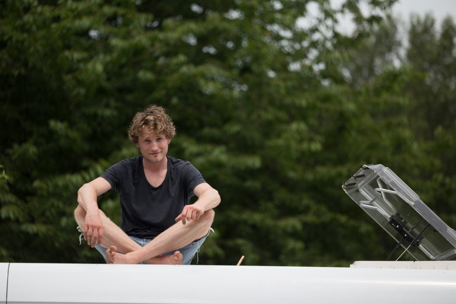 Jonge man met krullend haar zit in kleermakerszit op een wit camperdak voor groene bomen.