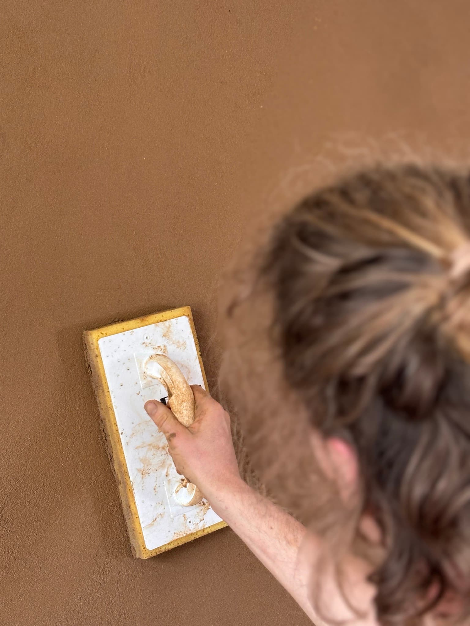 Een persoon strijkt een bruine, getextureerde muur glad met een wit schuurbord met spons.