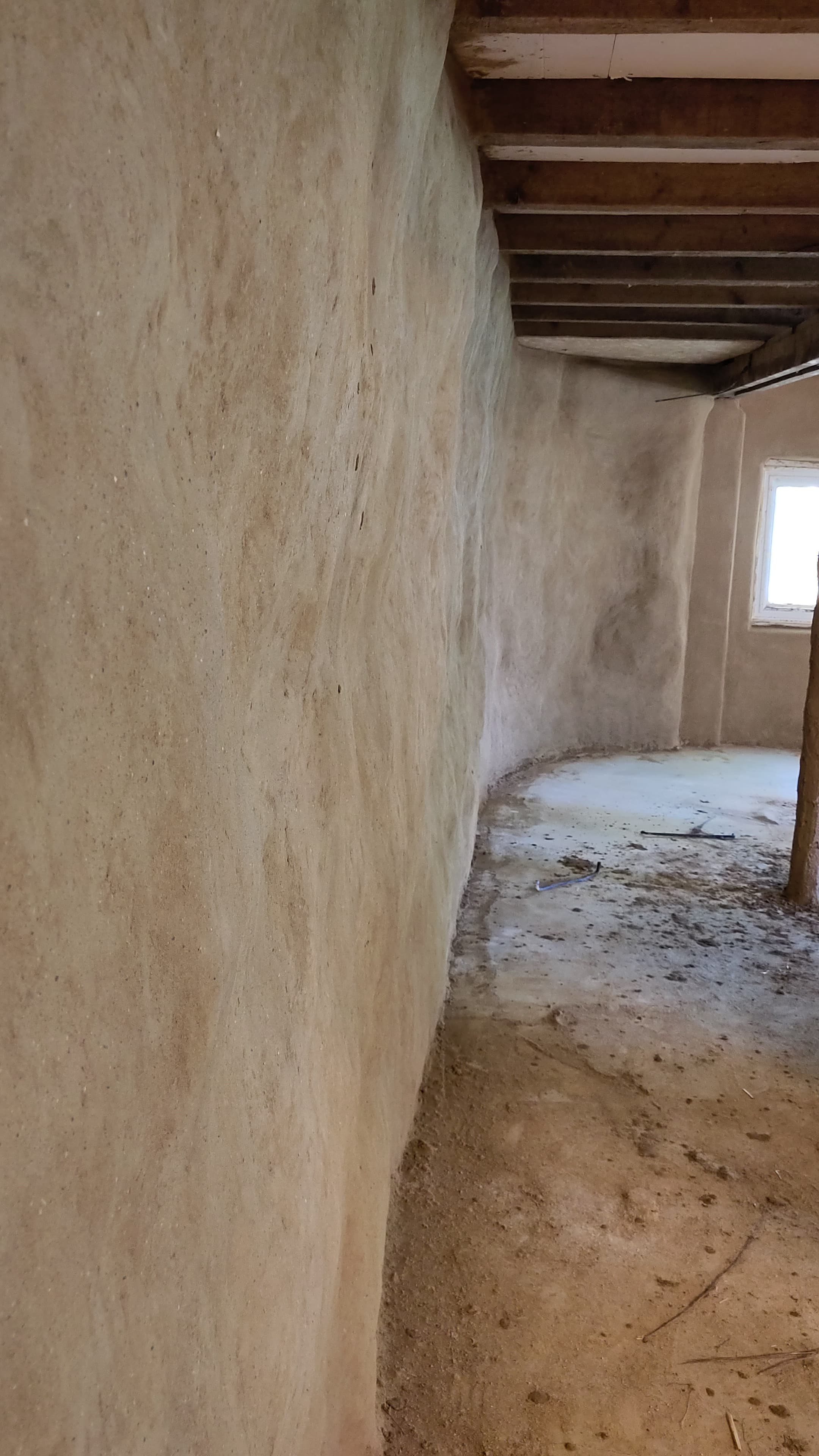 Een gebogen muur van getextureerd leemstuc in een kamer met zichtbare houten plafondbalken.