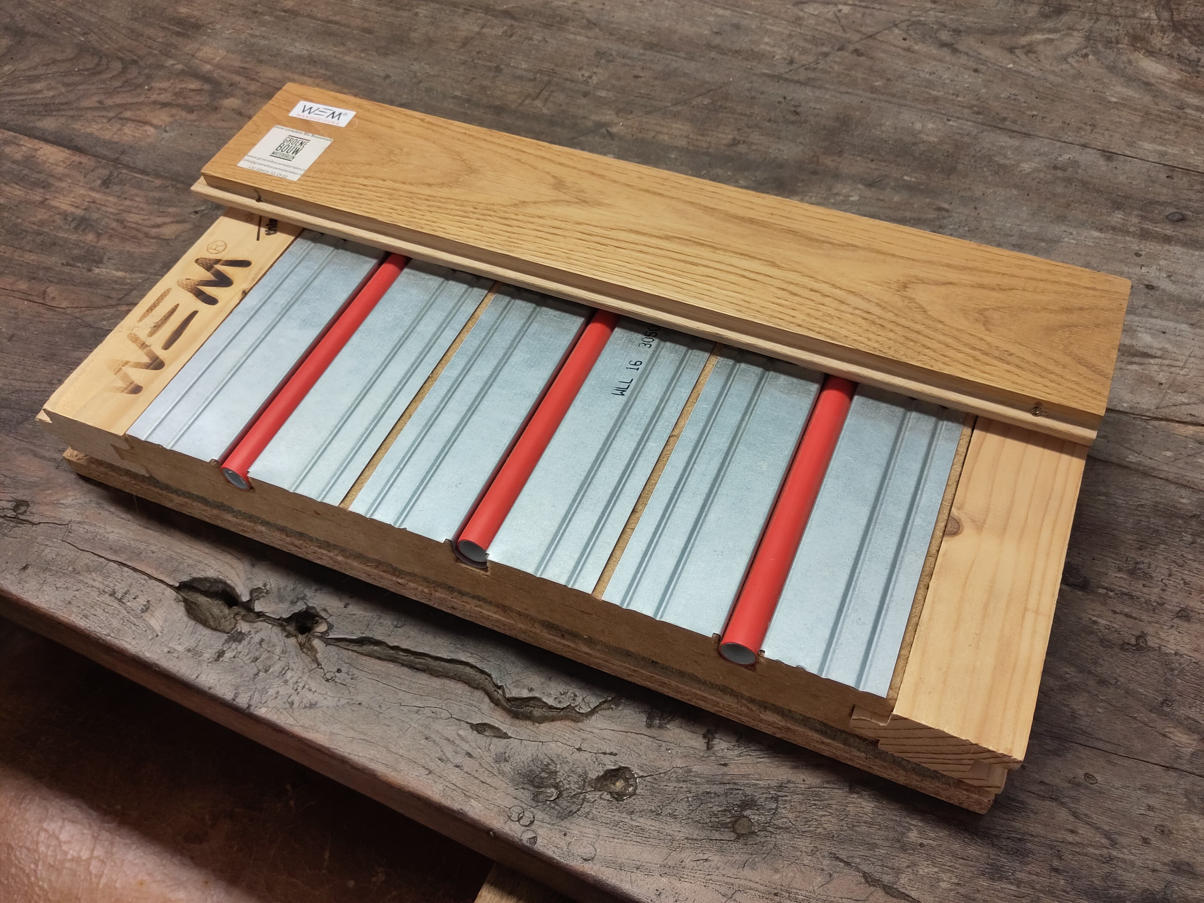 Doorsnede van een vloerverwarmingspaneel met rode buizen in metalen profielen onder een houten plank.
