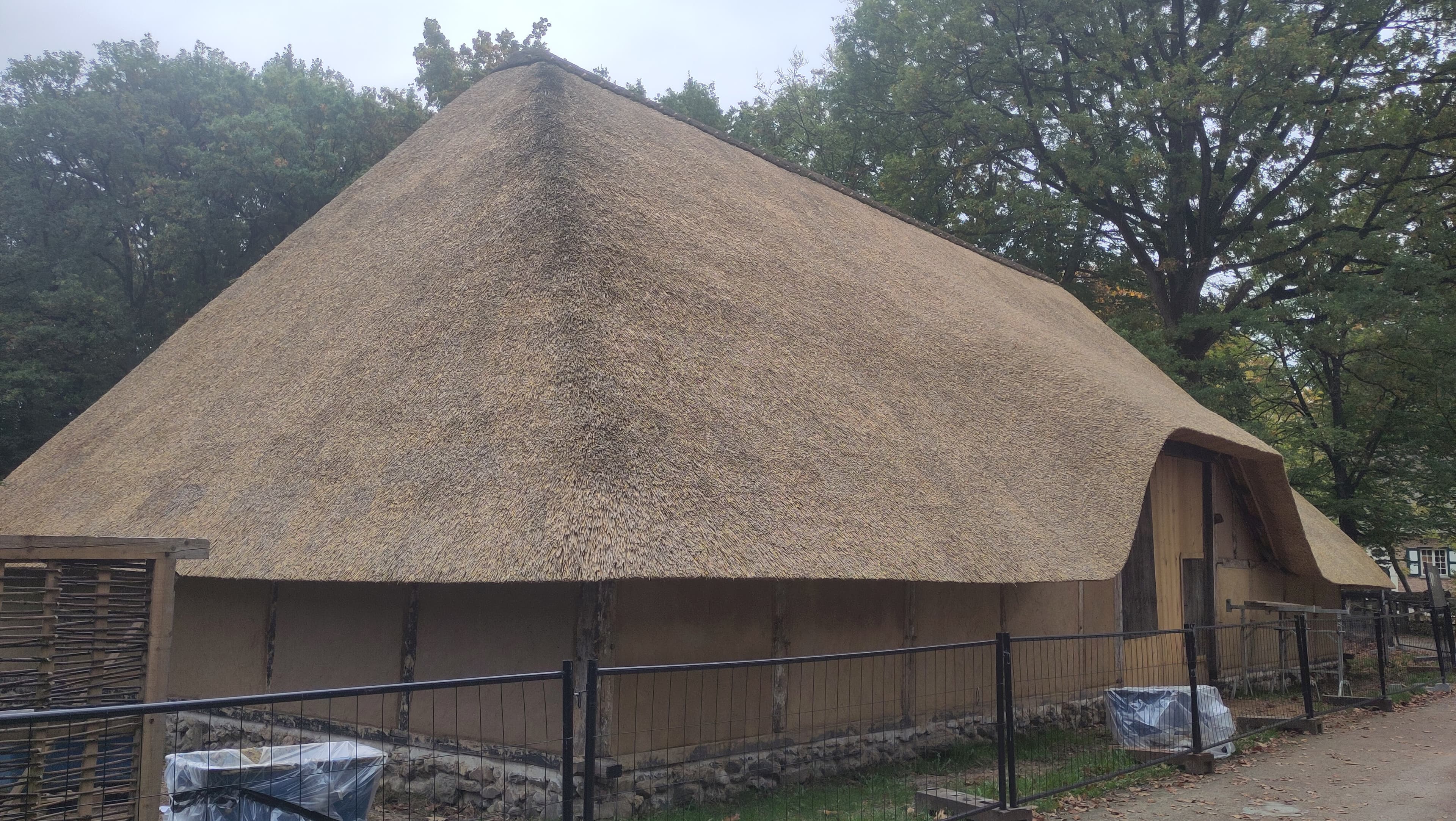 Historisch gebouw met een enorm rieten dak en vakwerkmuren, omgeven door bomen achter een hek.