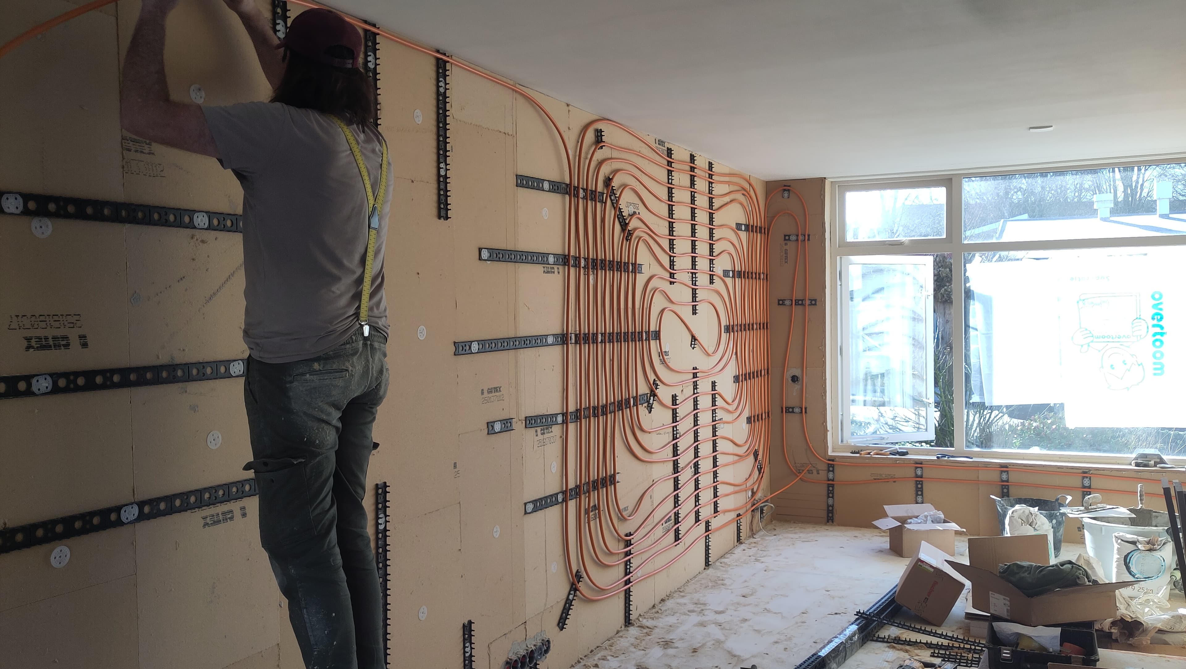 Een man bevestigt oranje verwarmingsbuizen in een spiraalpatroon op een muur tijdens renovatiewerkzaamheden.