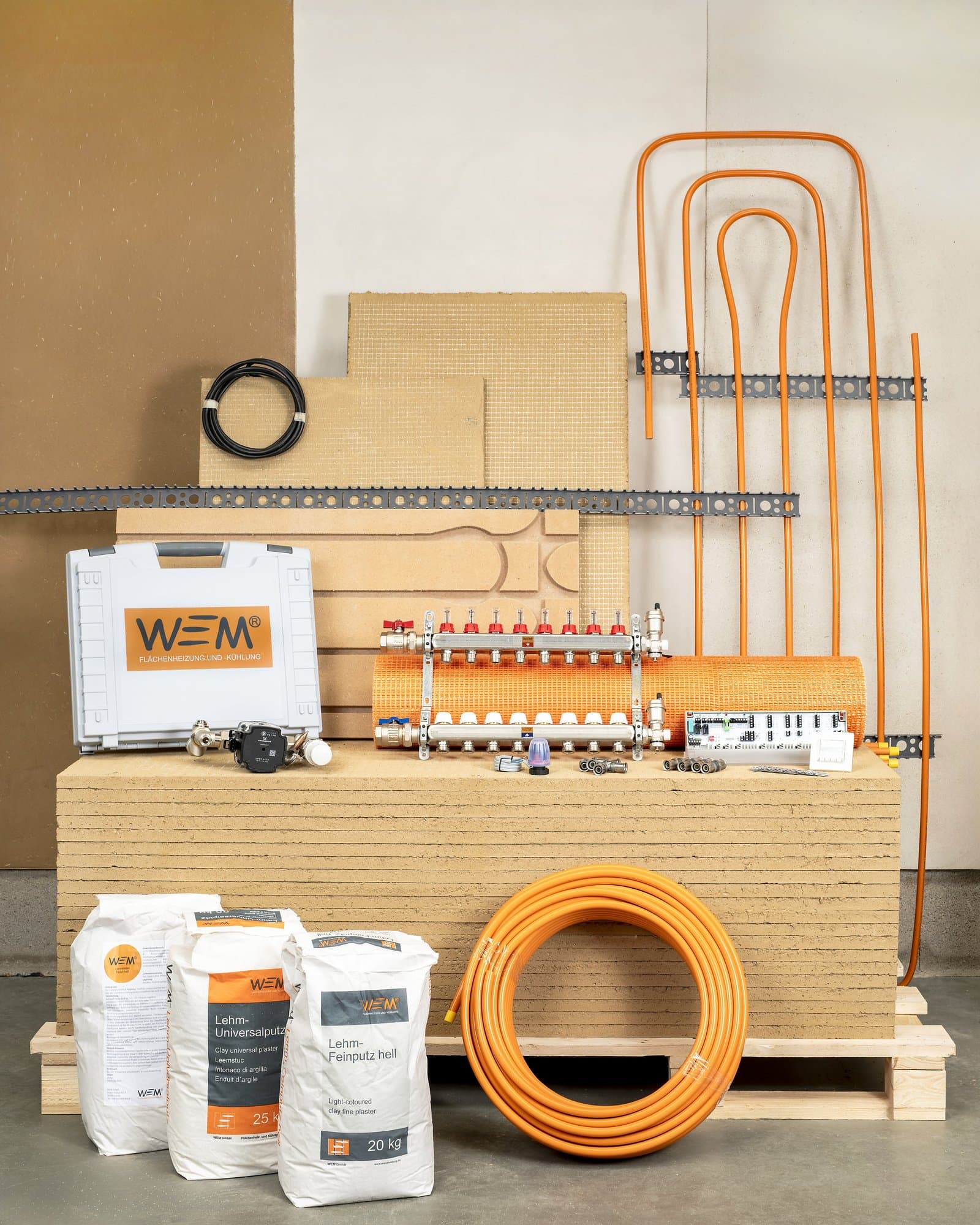 Verzameling WEM-materialen voor wandverwarming zoals leemplaten, oranje buizen, zakken leemstuc en een verdeler.