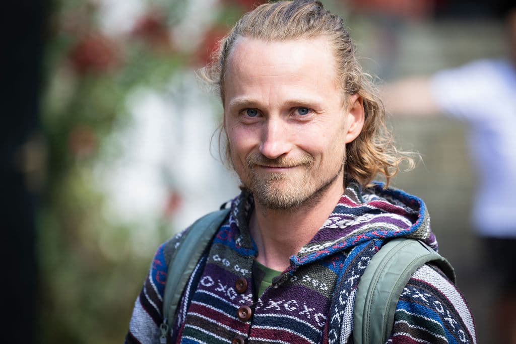 Glimlachende blonde man met een baardje, gekleed in een kleurrijke hoodie met patronen en rugzak.